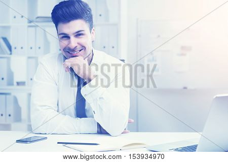 Young businessman in white shirt sitting at his office desk smiling to camera. Concept of corporate business and happiness. Toned image