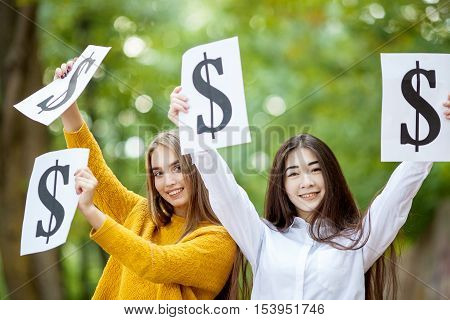Two young beautiful happy girl Asian and Europeans are holding the dollar symbol in the park summer day
