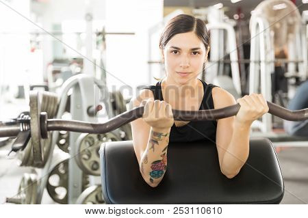 Portrait Of Confident Female Athlete Lifting Barbell Curl On Exercise Machine In Gym