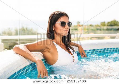 Picture of caucasian gorgeous woman in white bathing suit and sunglasses sitting in swimming pool at luxury hotel zone during vacation