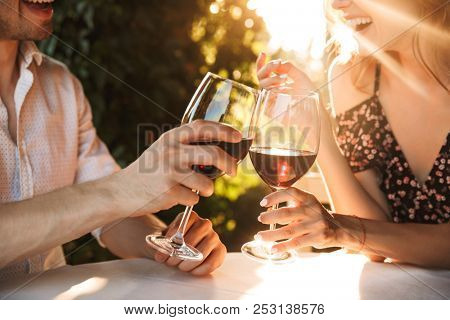 Cropped image of young loving couple sitting in cafe by dating outdors in park holding glasses of wine drinking.