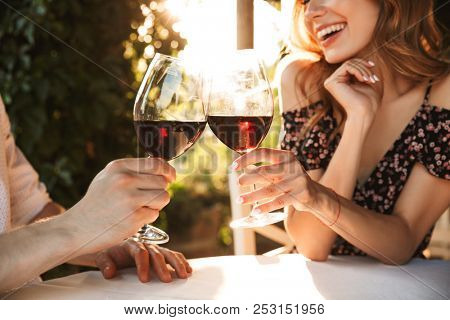 Cropped image of young loving couple sitting in cafe by dating outdors in park holding glasses of wine drinking.