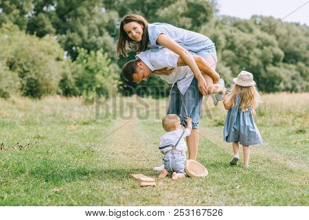 Happy Family With Two Kids Spending Time On Summer Feild, Father Piggybacking Mom