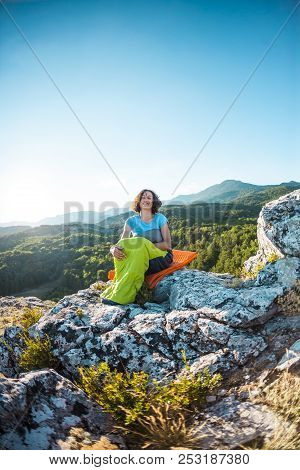 A Woman Is Sitting In A Sleeping Bag On Top Of A Mountain. A Smiling Girl Rests After A Climb. The B
