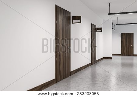 Hotel Corridor With White Walls, A Concrete Floor And A Row Of Closed Wooden Doors With Door Signs. 