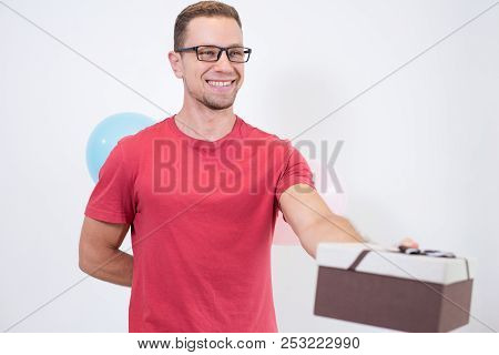 Cheerful Man Hiding Balloons Behind Back And Giving Gift Box. Smiling Handsome Young Delivery Guy In