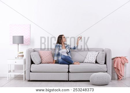 Young Woman Switching On Air Conditioner While Sitting On Sofa Near White Wall