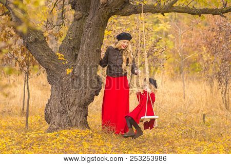 Beautiful Mother In A Long Dress Is Swinging On A Hinged Swing Little Daughter In A Red Coat In An A