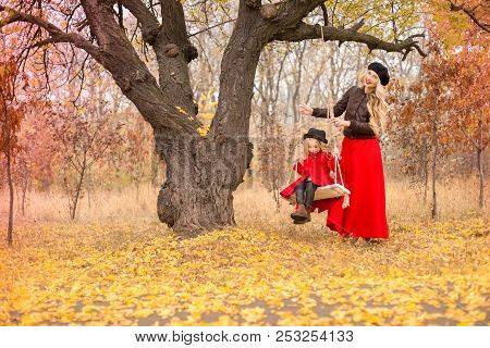 Beautiful Mother In A Long Dress Is Swinging On A Hinged Swing Little Daughter In A Red Coat In An A