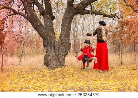 Beautiful Mother In A Long Dress Is Swinging On A Hinged Swing Little Daughter In A Red Coat In An A