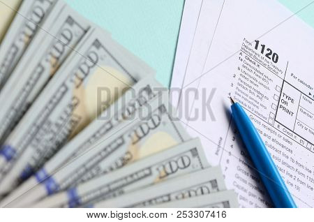 1120 Tax Form Lies Near Hundred Dollar Bills And Blue Pen On A Light Blue Background. Us Corporation