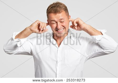Isolated Shot Of Displeased Male Pluggs Ears, Clenches Teeth, Ignores Loud Sound, Dressed In White E