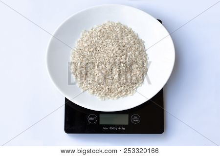 Raw Italian Rice In A White Plate On A Black Weight Scale On A White Background.