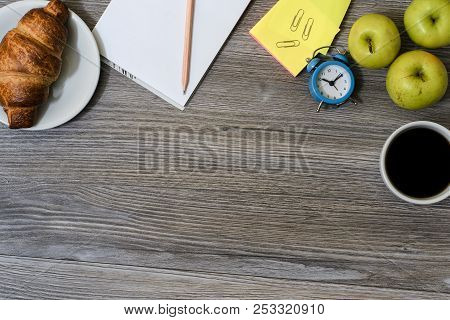 Close Up Of Workplace: Cup Of Coffee, Apples, Plate With Croissant, Notepad And Pencil, Alarm Clock 
