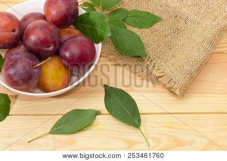 Fragment Of The White Bowl With Ripe Purple Plums And Leaves Of Plum Tree On A Light Colored Wooden 
