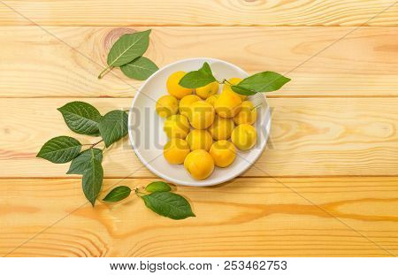 Yellow Cherry Plums On A White Saucer And Leaves On A Light Colored Wooden Surface