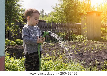 A Child In The Garden Watering Flowers, A Little Boy With A Water Hose Watering The Garden