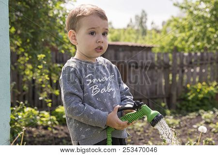 A Child In The Garden Watering Flowers, A Little Boy With A Water Hose Watering The Garden