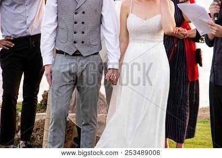 Happy Bride In White With Stones Dress And Bridegroom In Gray Fashionable Suit Holding Hands