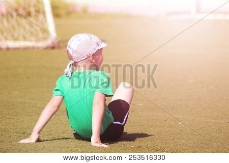 A Little Boy In A Green T-shirt Sitting On The Playing Football Field, Back View.