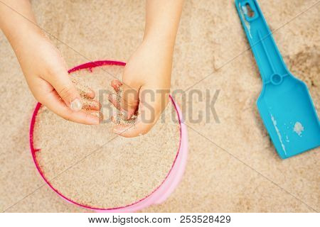 Hands Of A Girl With Sand Over A Colored Bucket And Shovel On The Beach