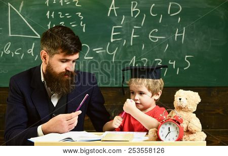 Hometask Concept. Teacher In Formal Wear And Pupil In Mortarboard In Classroom, Chalkboard On Backgr