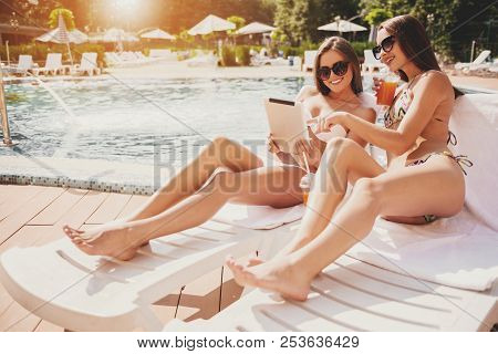 Two Beautiful Women Lying Using Tablet Near Pool. Digtal Device. Girls In Sunglasses. Rest Concepts.