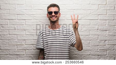 Young adult man wearing sunglasses standing over white brick wall showing and pointing up with fingers number three while smiling confident and happy.
