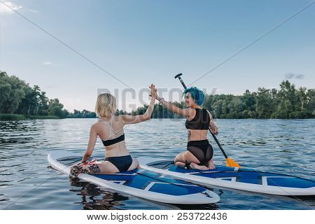 Athletic Women Giving Highfive While Sitting On Paddle Boards On River