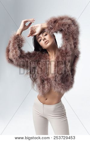 Young Elegant Girl Posing In Fluffy Pink Cape With Artificial Fur And Beige Flare Trousers On Gray B