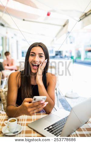 Young Latin Woman Getting Shocked By News In Her Phone Outdoors In Cafe. Shocked Face Of A Girl Sitt