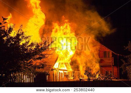 Burning Wooden House At Night. Bright Orange Flames And Dense Smoke From Under The Tiled Roof On Dar