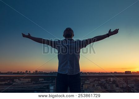 Man's Silhouette With Hands Rised Up To Sky On Sunset Cityscape Bacground. Feeling And Celebrating F