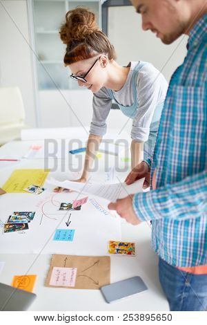 Two colleage students standing by desk while leaning over papers, pictures and notes and discussing them