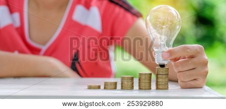 Hand Holding Light Bulb With Coins Stack On Wooden Table In The Morning. Energy And Money Saving , A