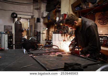 Serious concentrated young worker in leather jacket sanding metal part while producing metal frame in smithery