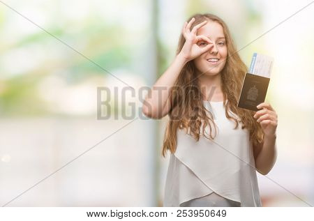 Young blonde woman holding passport of canada and boarding pass with happy face smiling doing ok sign with hand on eye looking through fingers