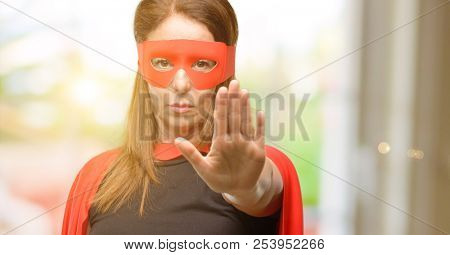 Middle age super hero woman wearing red mask and cape annoyed with bad attitude making stop sign with hand, saying no, expressing security, defense or restriction, maybe pushing