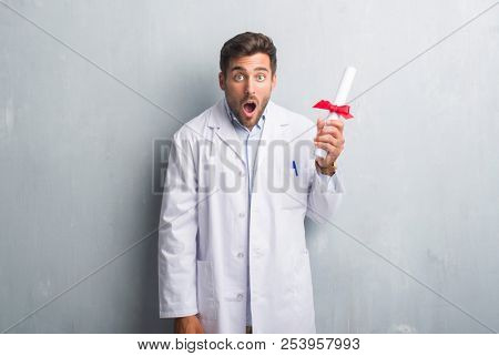 Handsome young doctor man over grey grunge wall holding diploma scared in shock with a surprise face, afraid and excited with fear expression