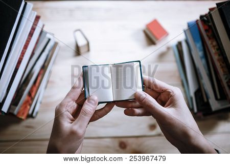 A Man Reading A Miniature Book Among The Big Books In The Library At The Wooden Table.