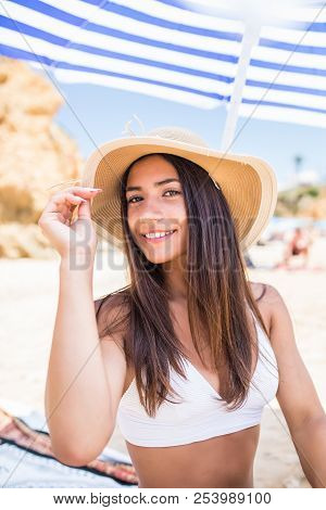Young Beauty Latin Woman In Bikini And Straw Hat Sitting Under Sun Umbrella On The Beach Near Sea Co