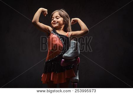 Cute Little Girl Dressed In Skater Dress Holds Ice Skates And Shows Muscles. Isolated On Dark Textur