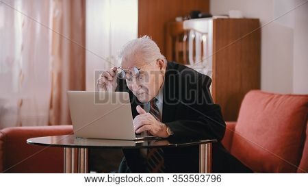 An Old Businessman In Glasses Sitting By The Laptop At Looking At The Monitor