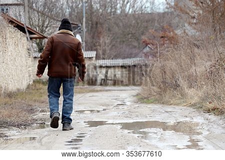 Rain In A Countryside, Man In Jeans And Dirty Galoshes Walks On A Rural Street With Puddles. Rainy W
