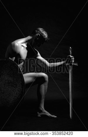 Side View Of Strong Warrior Standing On Knee And Looking Down. Monochrome Portrait Of Muscular Athle