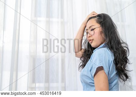 An Asian Glasses Woman In Blue Shirt  Poses By Slicks Her Hair Back In Front Of Lightening Curtain.