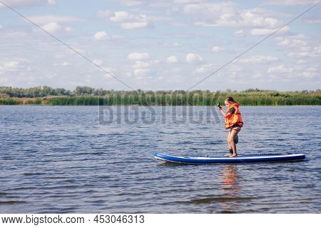 Small Girl Paddle Boarding By Herself On Lake With Oar In Hands In Orange Life Jacket. Active Holida