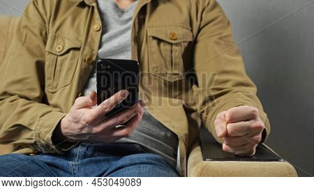 Man Gets Angry After Losing Lottery Hitting Armrest With Fist Sitting In Armchair Against Grey Wall.