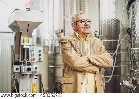 Portrait Of Happy Senior Man Who Owns Winery. He Is Standing Beside Wine Storage Tanks. Industry Win