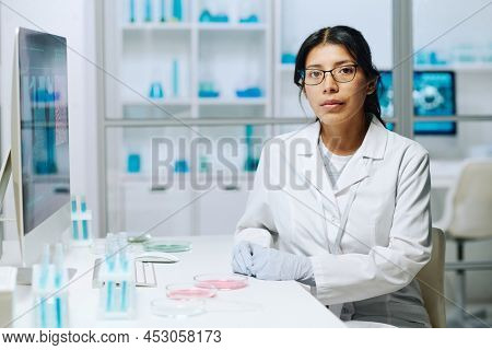 Young serious female virologist or microbiologist in whitecoat looking at camera while sittiing by workplace with test tubes and petri dishes